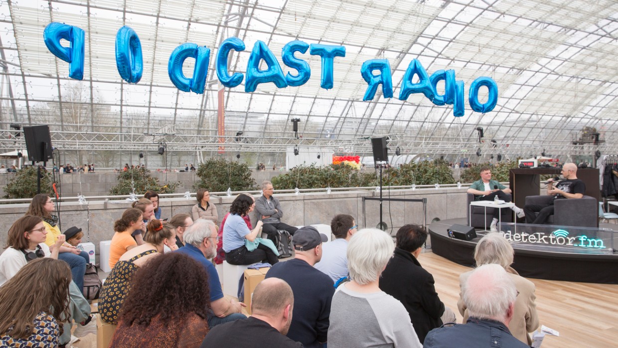 Auf der Lesebühne von detektor.fm findet eine Lesung vor Publikum in der Glashalle während der Leipziger Buchmesse 2024 statt. Darüber schweben blaue Luftballons mit dem Schriftzug "Podcast Radio".