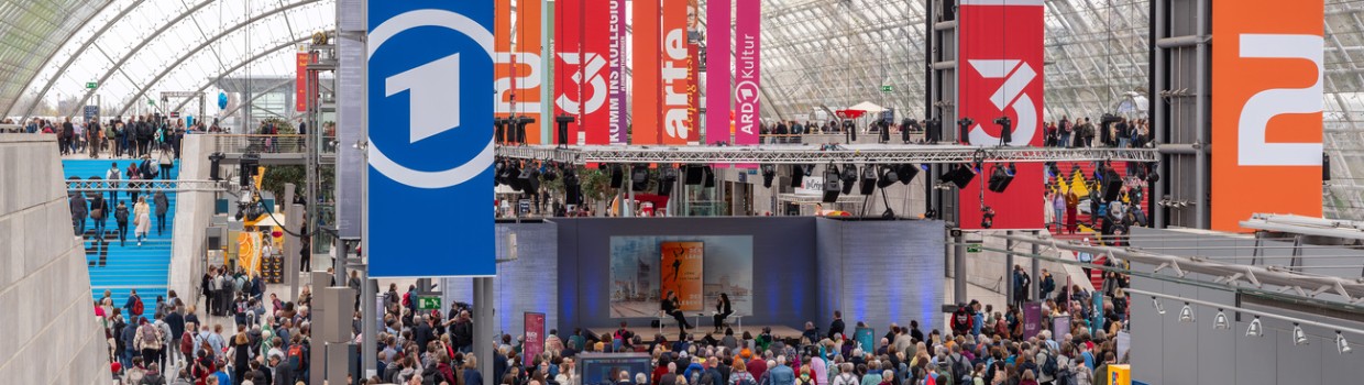 Innenaufnahme der Glashalle der Leipziger Messe mit bunten Werbebannern und vielen Besucher:innen während der Leipziger Buchmesse 2024