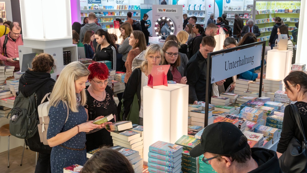Besucher:innen betrachten Bücherstapel auf der Leipziger Buchmesse 2024 in der vollen Messehalle