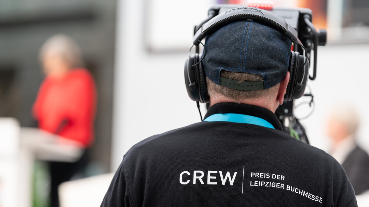 Kameramann von hinten mit großen Kopfhörern und einem T-Shirt mit der Aufschrift "Crew", der mit einer großen Kamera die Preisverleihung während der Leipziger Buchmesse 2024 filmt