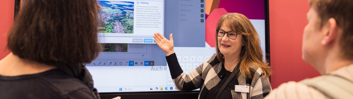 Ausstellerin im Bildungsbereich stellt auf der Leipziger Buchmesse 2025 mit Hilfe einer digitalen Wand zwei anderen Frauen ein Lernprogramm vor Ausstellerin im Bildungsbereich stellt auf der Leipziger Buchmesse 2025 mit Hilfe einer digitalen Wand zwei anderen Frauen ein Lernprogramm vor